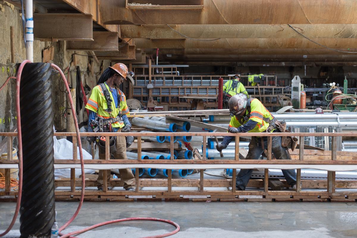 Construction workers in safety vests assemble wooden formwork at an underground transit station platform.