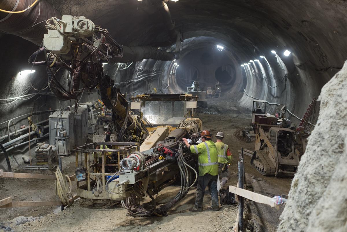 Workers in safety vests maintain drilling equipment inside a large underground tunnel excavation site.