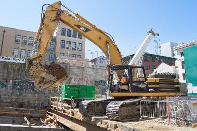 Yellow excavator removing soil from headhouse exterior on urban construction site with green debris container and crane nearby.