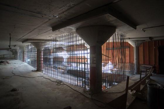 Under-construction transit station interior with concrete pillars, metal rebar framework, and orange safety barriers.