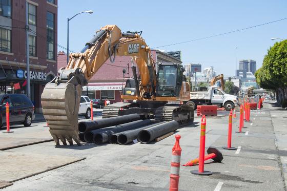 Large excavator loading black sewer pipes onto a truck on a city street with traffic cones and urban buildings visible.
