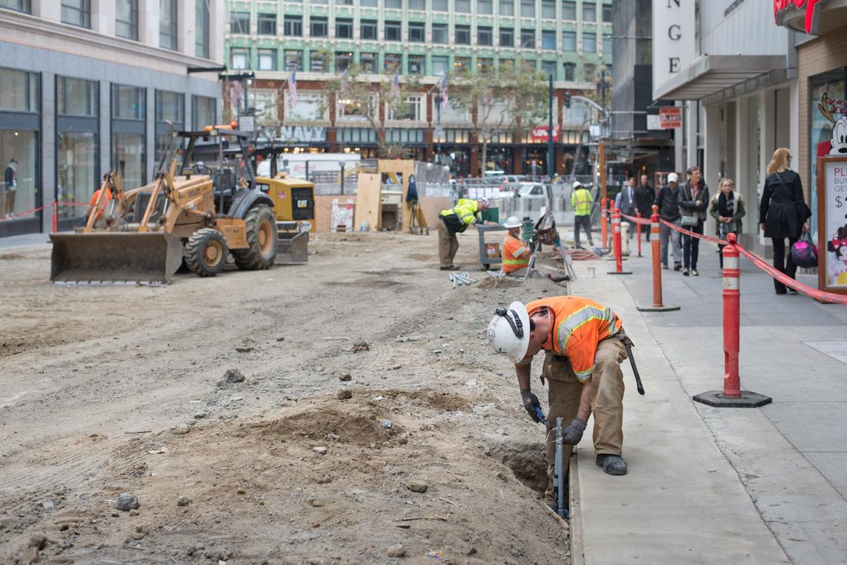 Construction workers in safety gear perform underground infrastructure work on a city street with heavy equipment and temporary barriers.