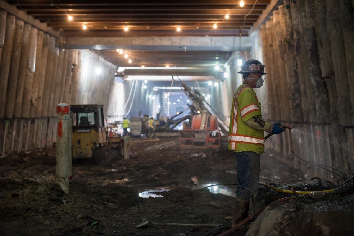 Worker in safety vest and hard hat inside underground transit tunnel with construction equipment and lighting.