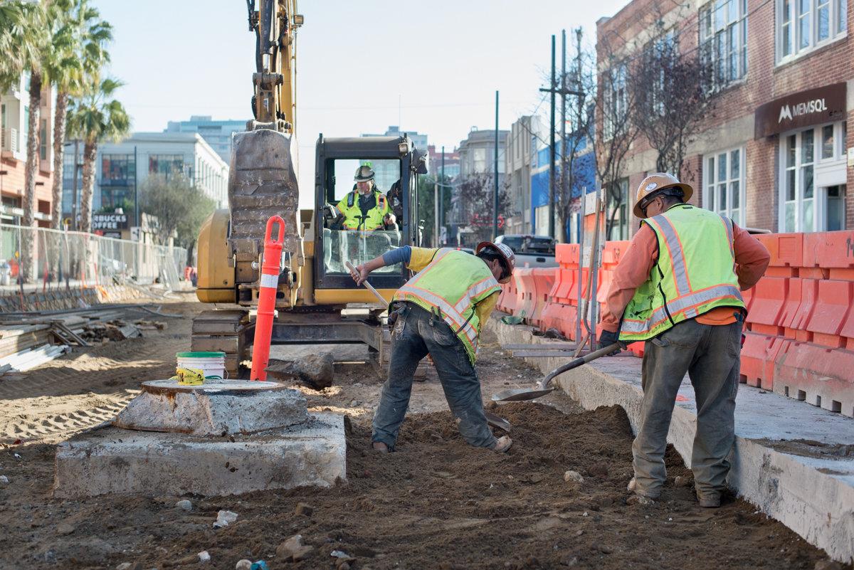 Construction workers in safety vests prepare ground for track installation using hand tools and excavator equipment on urban street.