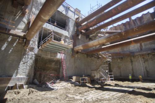 Construction site showing concrete pillars, steel beams, and excavation work inside a station box with temporary bracing structures.