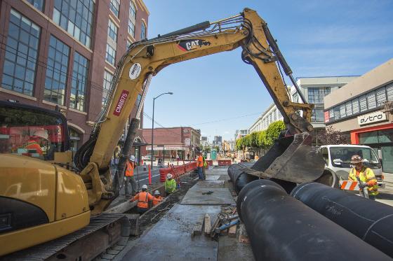 Yellow excavator installing large black duct bank in street trench with construction workers and equipment nearby.