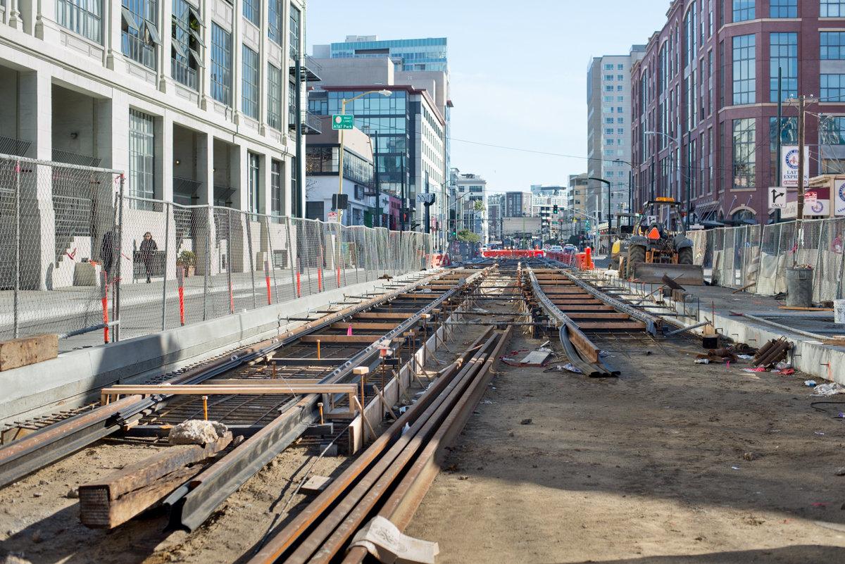 Construction site showing streetcar track installation on 4th Street with metal rails, fencing, and urban buildings.