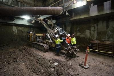 Workers in safety vests operate drilling equipment in underground station construction area with steel support beams.