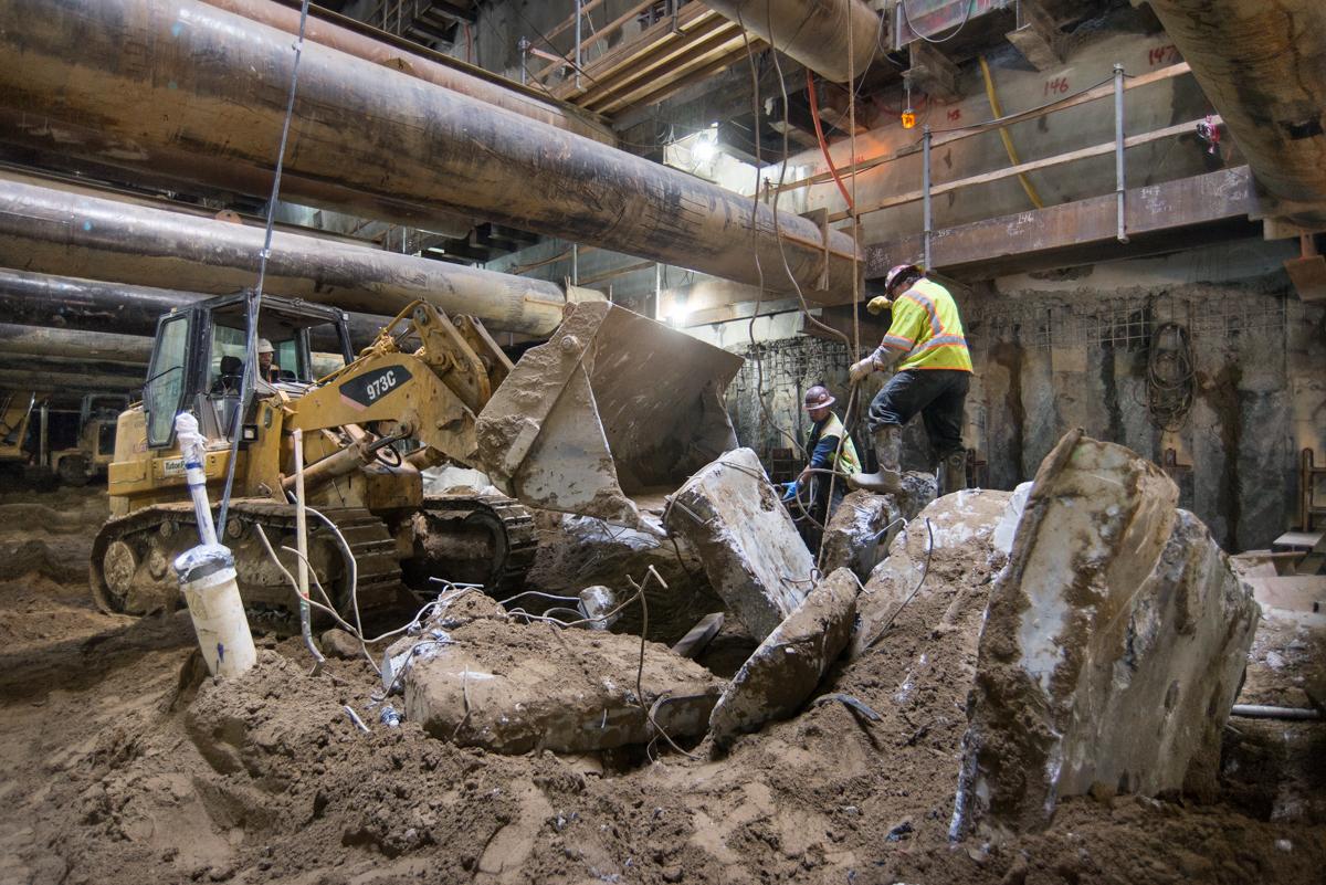 Construction crew uses bulldozer to remove demolished tunnel ring segments from underground station excavation site.
