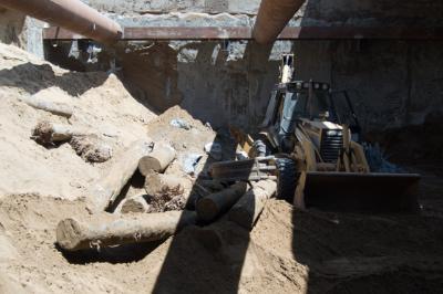 Construction excavation site with temporary steel support structures and exposed wooden pilings beneath concrete structure.
