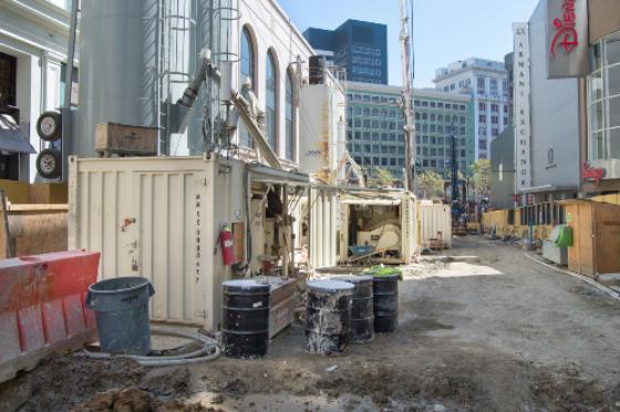 Construction site with shipping containers and equipment on urban street surrounded by buildings.