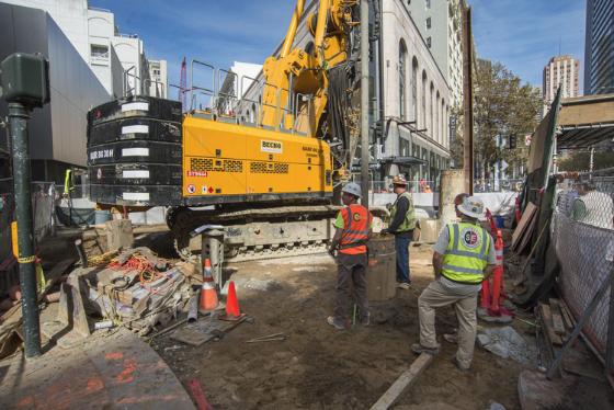 Construction crew operates yellow excavator at Union Square/Market Street Station work site with traffic cones and safety barriers visible.