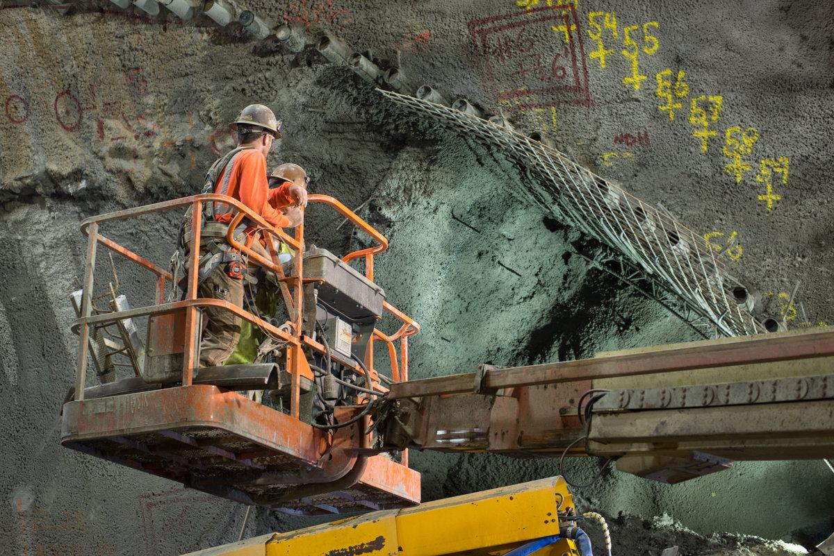 Worker in orange safety vest on elevated platform inspecting drill casings on concrete wall at transit construction site.