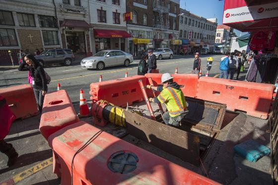 Worker in safety vest inspects waterline replacement work in street excavation at Stockton and Washington streets.