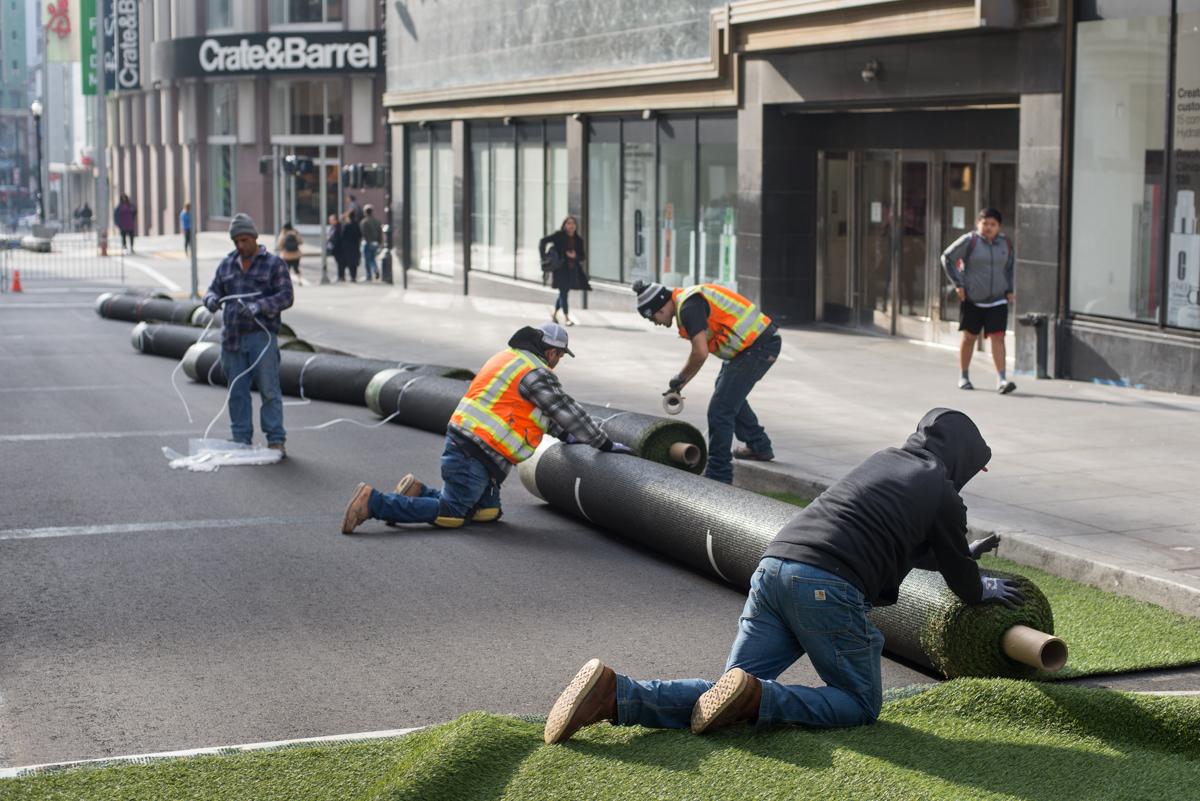 Workers in safety vests rolling up green turf segments on a downtown street near Crate and Barrel.
