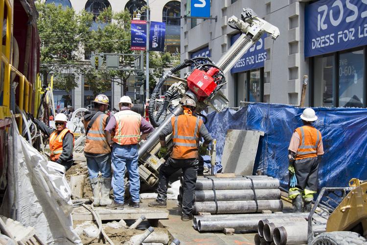 Construction workers in safety vests operate drilling equipment on a downtown street surrounded by blue protective barriers and pipes.