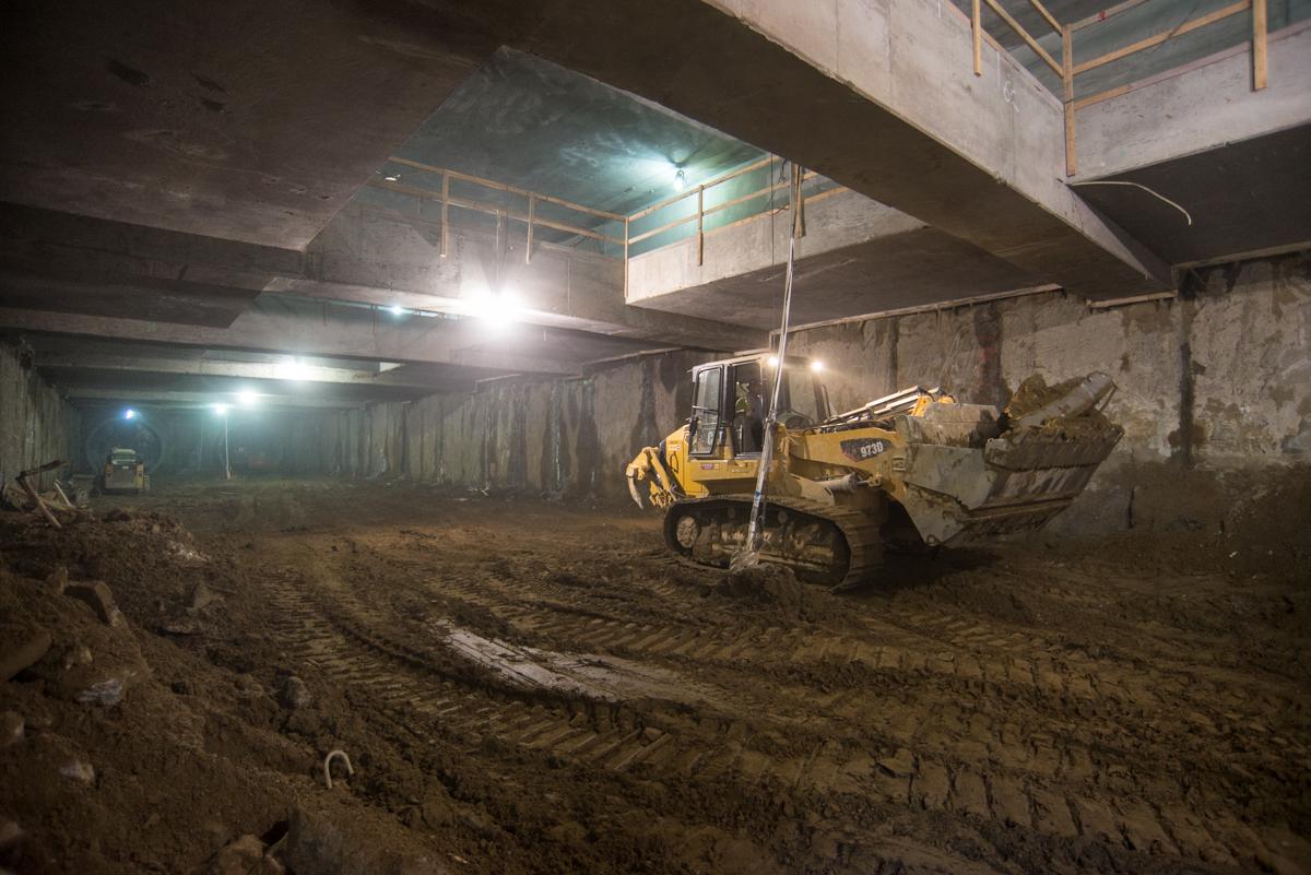 Bulldozer excavating underground station platform beneath concrete structure with temporary lighting.
