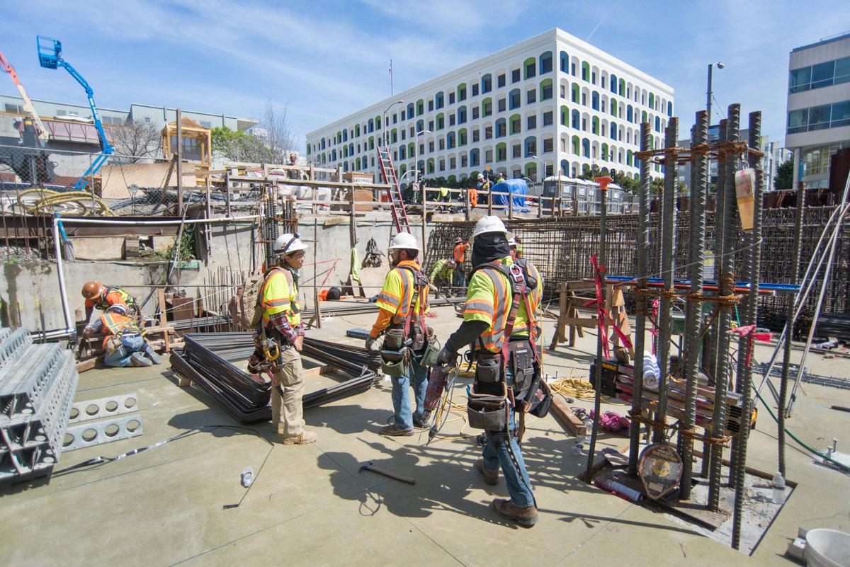 Construction workers in safety gear work on concrete foundation with rebar and steel framework at urban transit station site.
