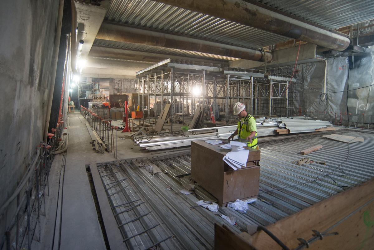 Worker reviewing plans on mezzanine level of underground station construction site with exposed structural framework and corrugated metal ceiling.