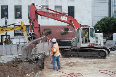 Red excavator with operator in safety vest sorting demolished concrete debris at construction site near building.