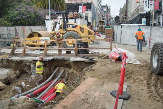 Construction crew working on underground utilities at 4th and Clementina Street with heavy equipment and exposed pipes.
