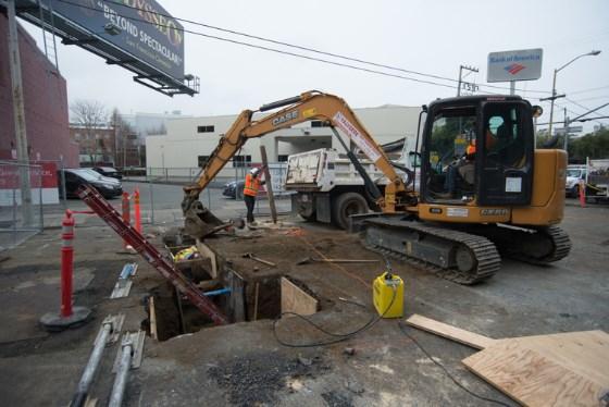 Construction crew operating excavator at underground utility work site with trench shields and safety barriers.