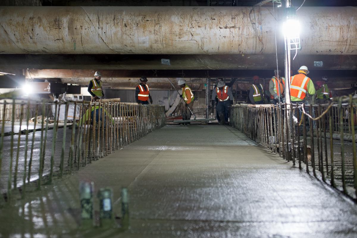 Construction workers in safety vests work on a wet concrete platform beneath a large pipe, with metal railings lining both sides.