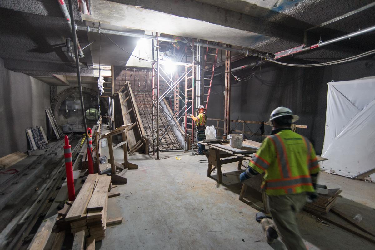 Construction workers in safety vests install scaffolding and concrete forms inside a transit station.