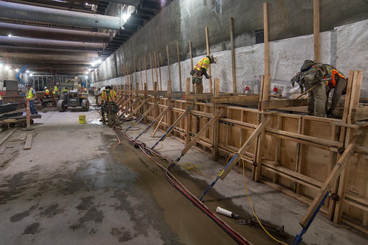 Construction workers install wooden forms and steel bracing inside underground transit station excavation.
