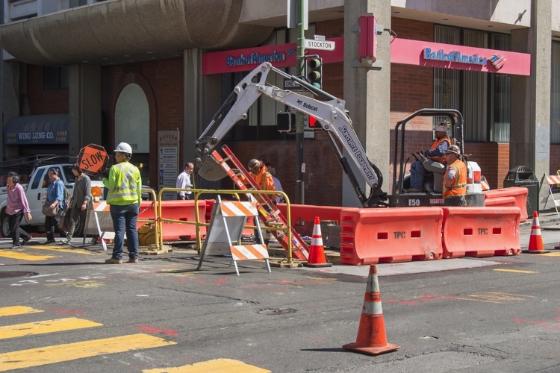 Construction crew operates excavator on street near Stockton and Washington, with red barriers and traffic cones marking work zone.