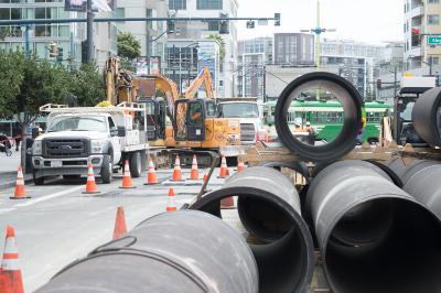 Large sewer pipes staged for installation during utility upgrade work on city street with construction equipment and traffic control.