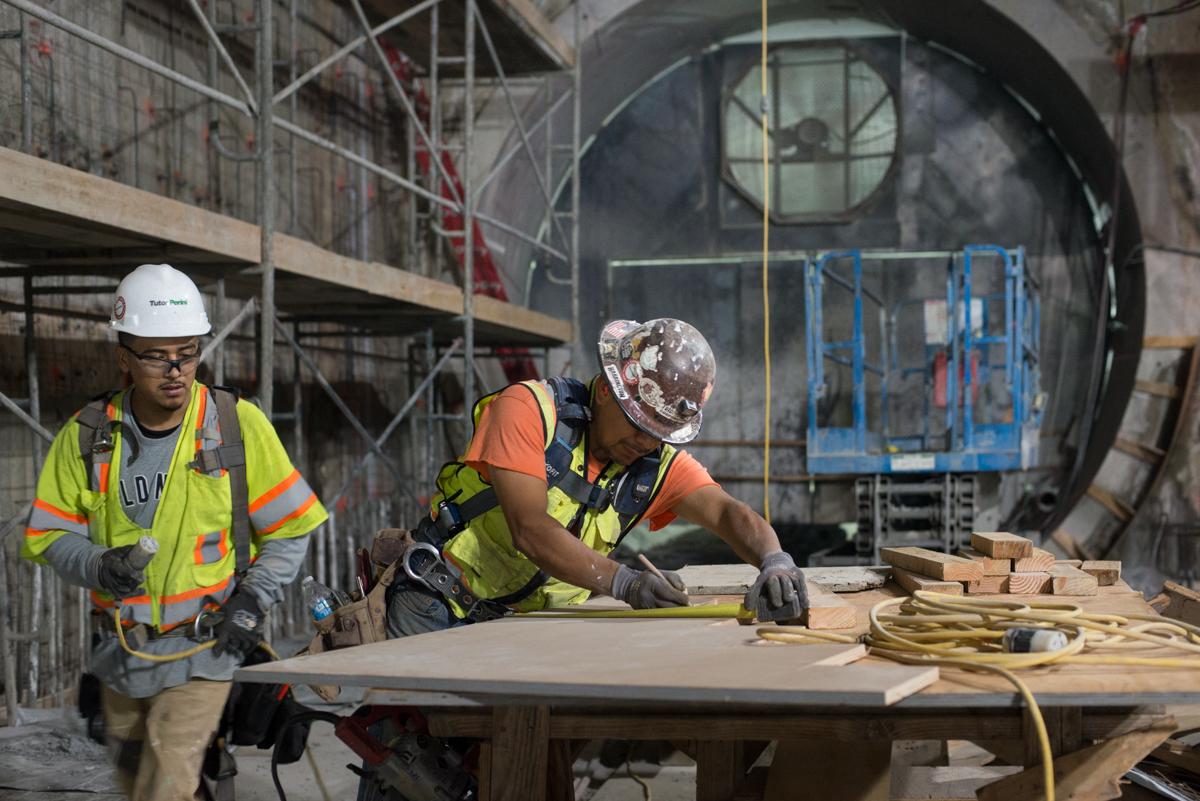 Two construction workers in safety gear work on concrete forms at a transit station construction site.