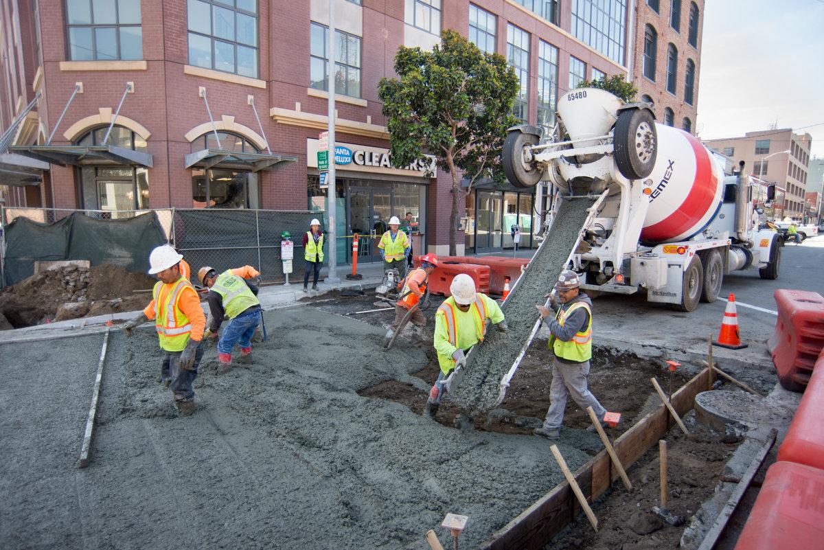 Construction workers guide concrete from mixer truck into street trench on urban street.