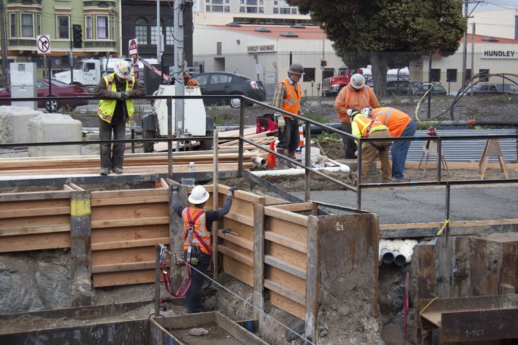 Construction workers in safety vests and hard hats work on a retaining wall at a transit construction site with wooden barriers and metal railings.