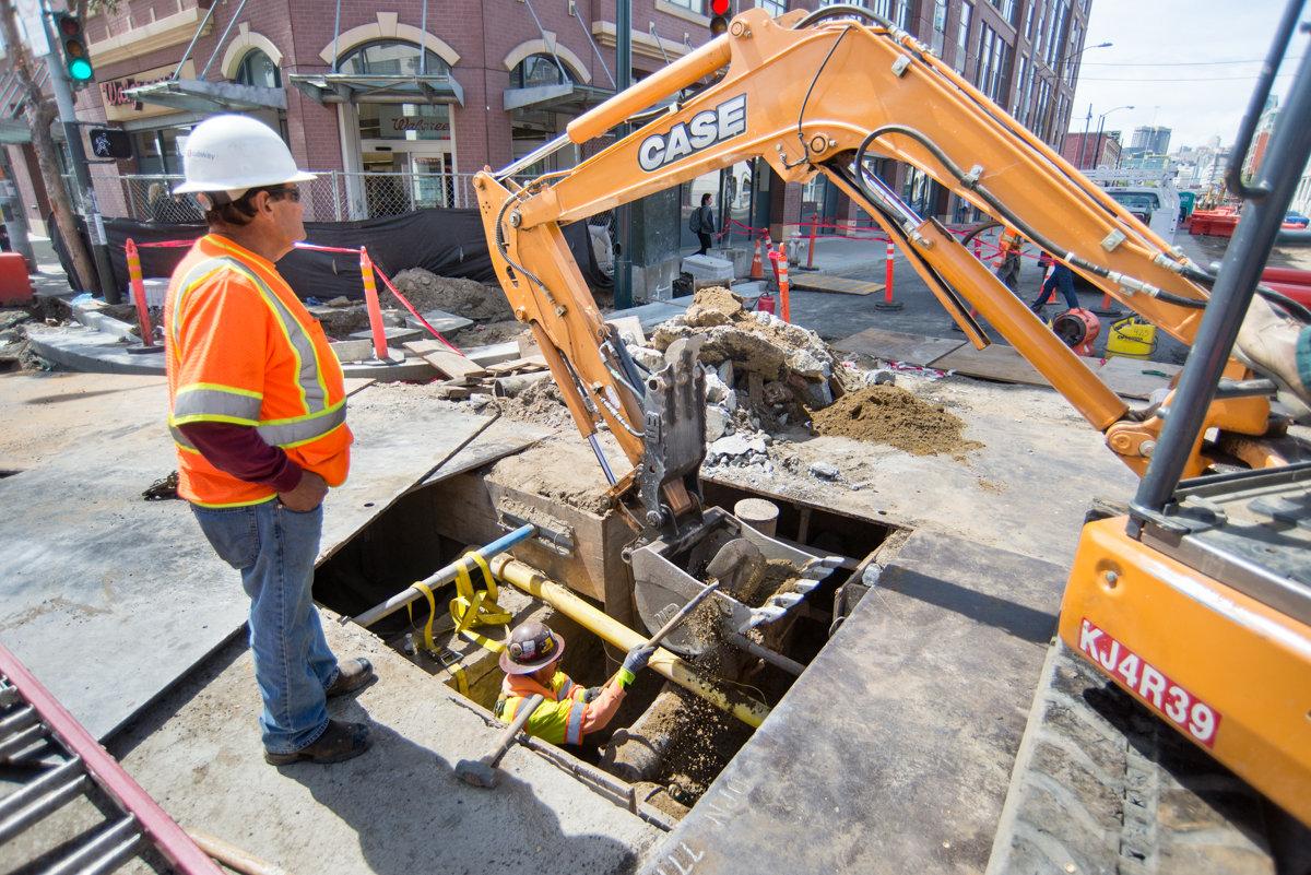 Construction worker in safety gear observing CASE excavator removing old utility infrastructure from street excavation pit.