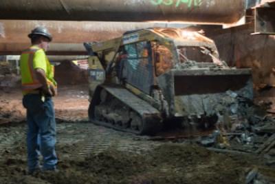 Worker in safety vest observing tracked excavator removing debris from construction site.