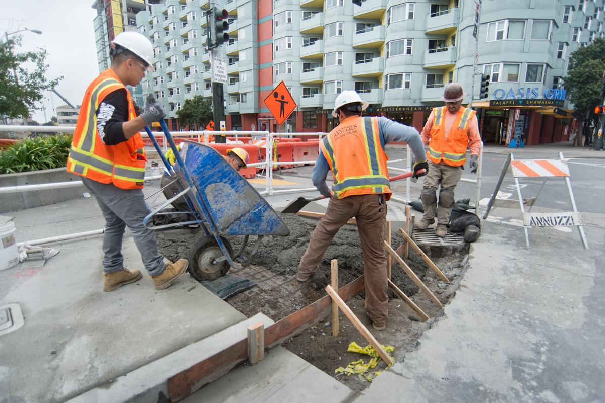 Construction workers in safety vests operate a wheelbarrow to move concrete debris at a street excavation site near apartment buildings.