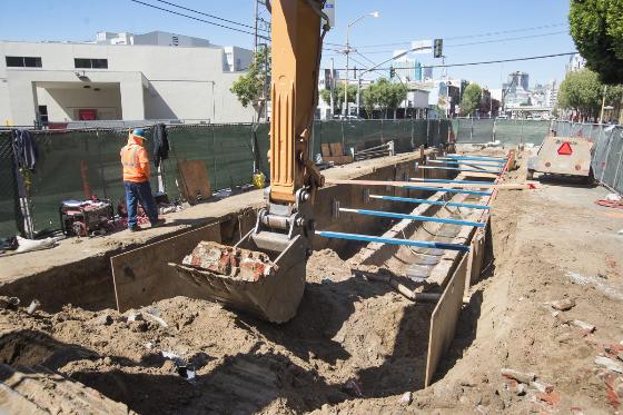 Excavated roadway with shoring installed around underground sewer line; construction equipment and worker visible.