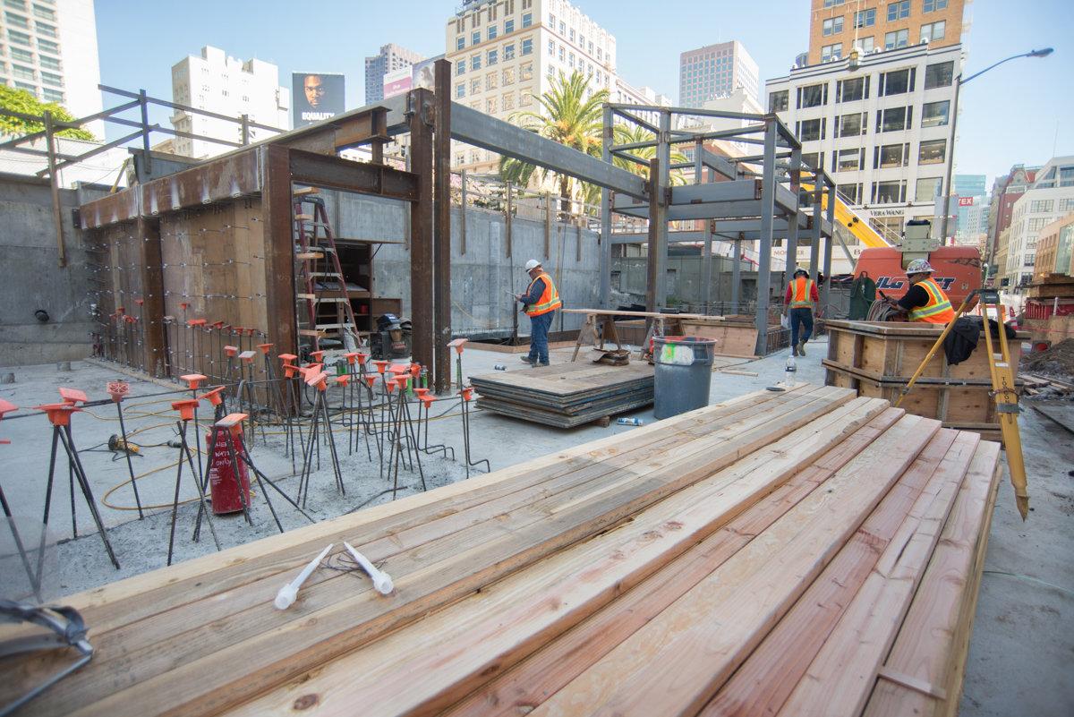 Construction site at 4th Street with workers, steel framework, wooden planks, and concrete foundation work in urban setting.