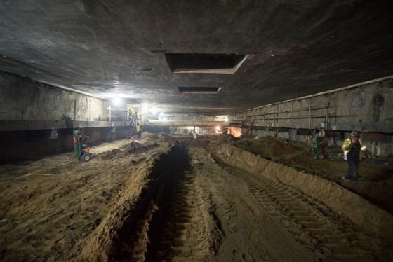 Underground station box construction with two parallel elevator shaft trenches and workers in hard hats.