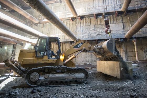 Excavator with hydraulic arm operating inside underground station construction site with concrete beams overhead.