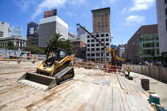 Construction site with yellow excavator on wooden platform, urban buildings in background, street closure barriers visible.