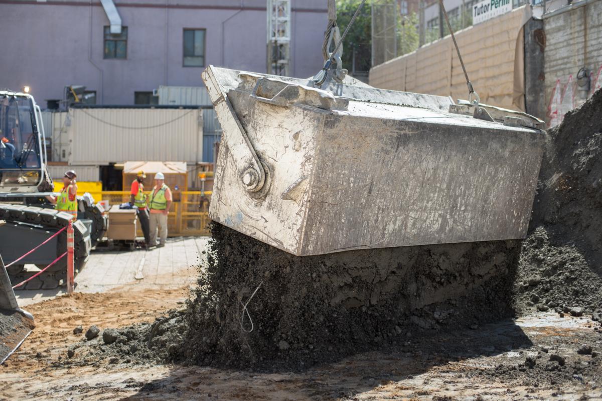 Large steel excavation bucket suspended by crane above construction site with workers and equipment in background.