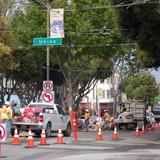 Street intersection with green transit sign, construction barriers, and trees during daytime.
