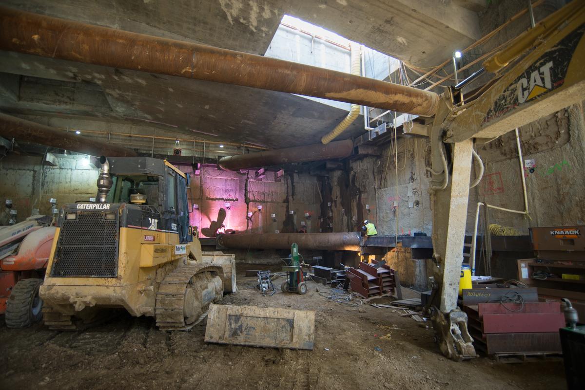 Industrial underground construction site with heavy equipment, steel supports, and pink safety barriers inside a transit station headhouse.