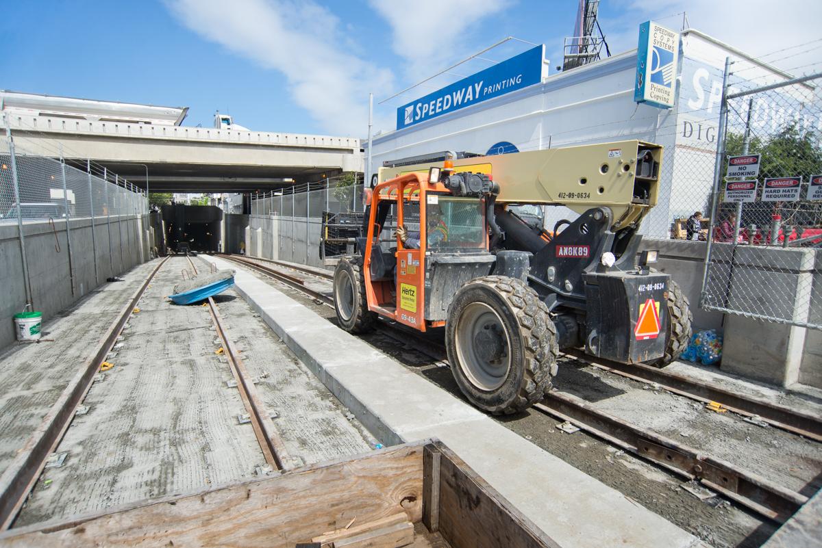 Orange telehandler lifting construction materials on 4th Street near tunnel portal, with concrete forms and track visible.
