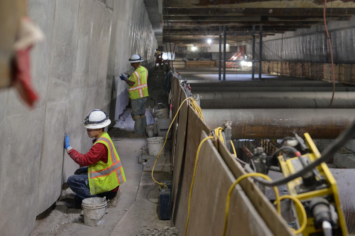 Workers in safety gear inspect concrete wall during underground transit construction.