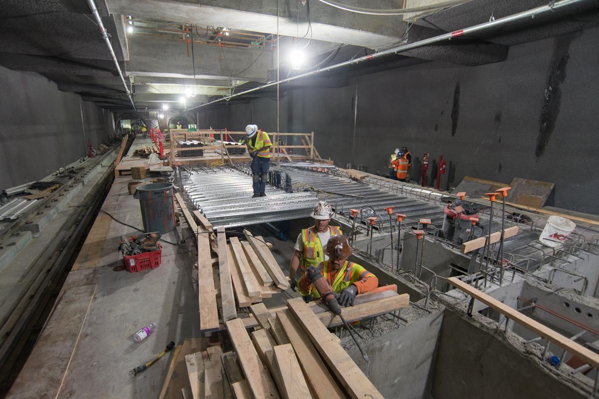Workers install steel decking and rebar for station platform construction in underground transit tunnel.