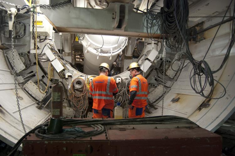 Two workers in orange safety vests and hard hats assemble tunnel boring machine components underground.