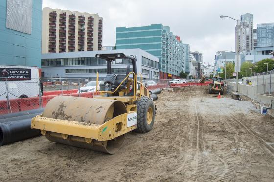 Road roller compacting soil on 4th Street construction site with downtown buildings in background